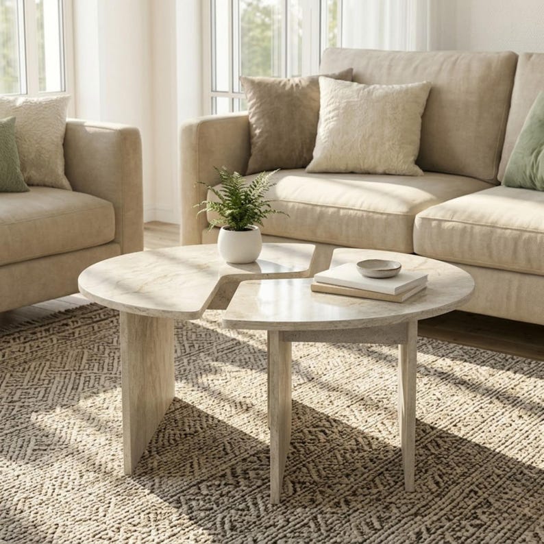 May include: A light-colored, round coffee table with a unique cutout design. The table has a light wood finish and is supported by three legs. A small potted plant and books are on the table, which sits on a patterned rug.