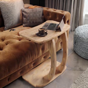 May include: A light-toned wooden C-shaped side table with a smooth rectangular top and a curved base. The table holds a silver laptop and a dark ceramic mug filled with coffee. The wood grain is visible throughout the structure of the furniture.