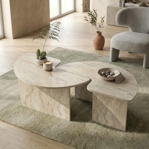 May include: A two-piece, light beige stone coffee table with a unique, curved design. The table sits on rectangular supports and is placed on a light green rug. Decorative items include a small cactus, a candle, and a bowl with stones.