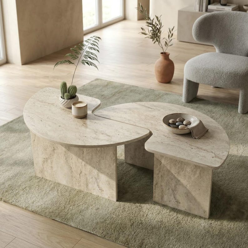 May include: A two-piece, light beige stone coffee table with a unique, curved design. The table sits on rectangular supports and is placed on a light green rug. Decorative items include a small cactus, a candle, and a bowl with stones.