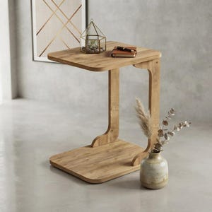 Può includere: Un tavolino a forma di C realizzato in legno di tonalità chiara con una finitura liscia. Il tavolo presenta un piano rettangolare piatto e una base abbinata collegati da un unico supporto verticale. Un terrario geometrico in vetro è appoggiato sopra.