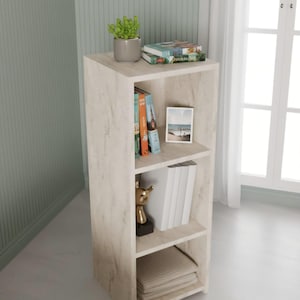 Puede incluir: Una estantería de tres estantes con un acabado claro similar al mármol. El estante superior contiene una pequeña planta en maceta, libros y una foto. El estante del medio muestra libros y una estatua dorada. El estante inferior tiene toallas dobladas.