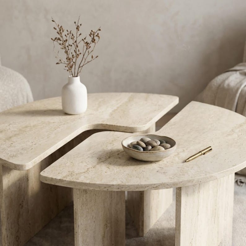 May include: Two-piece travertine coffee table set with a unique curved design. The tables feature a light beige color and are accessorized with a white vase, a small bowl of stones, and a gold pen. The tables are a modern addition to any living room.