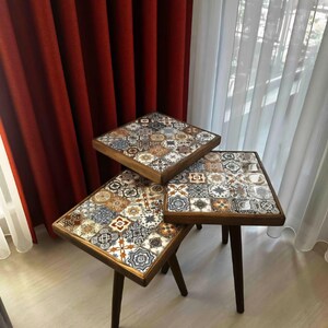 Op de afbeelding: Drie vierkante bijzettafels met donkere houten poten en frames. De tafelbladen hebben een mozaïektegelontwerp in tinten bruin, blauw en crème. De tafels zijn in een trapsgewijze opstelling gerangschikt.
