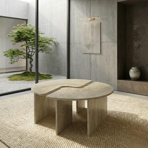May include: A modern, light-colored stone coffee table with a unique, split-circle design. The table sits on a beige woven rug. The room features a minimalist aesthetic with a tree visible through a large window.