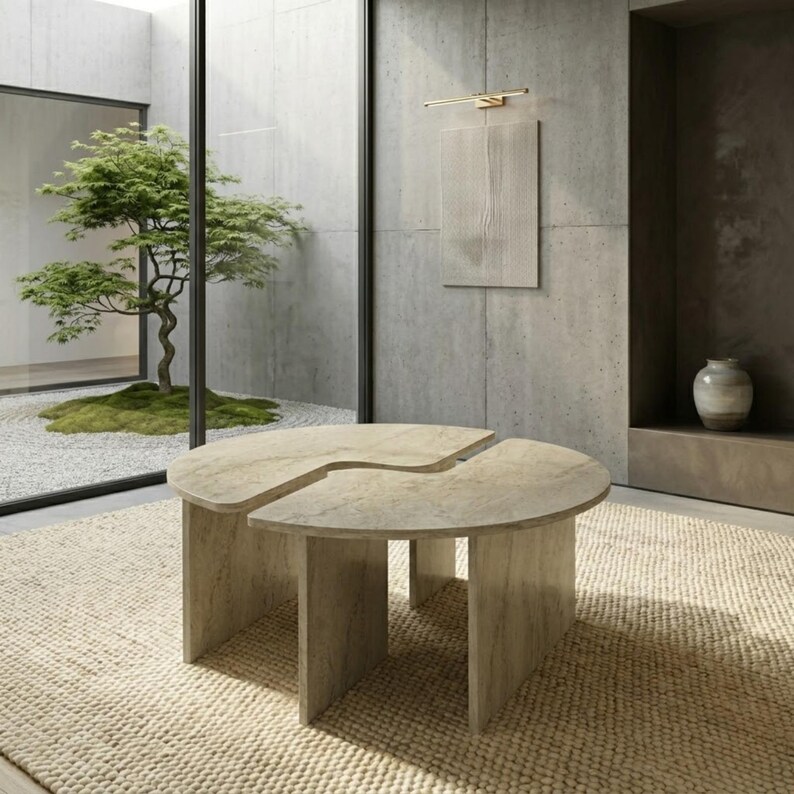 May include: A modern, light-colored stone coffee table with a unique, split-circle design. The table sits on a beige woven rug. The room features a minimalist aesthetic with a tree visible through a large window.