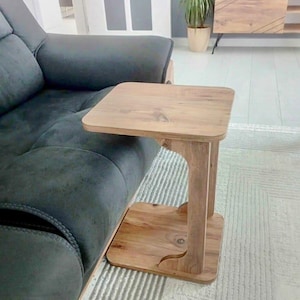 May include: A wooden side table with a square top and a lower shelf. The table is made of light-colored wood and has a curved design on the lower shelf. It is positioned next to a dark gray sofa.