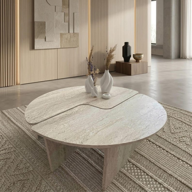 May include: A modern, light-colored stone coffee table with a unique, split-circle design. The table is supported by three angled legs and sits on a patterned rug. Two white vases with dried flowers and a small decorative object are on the table.