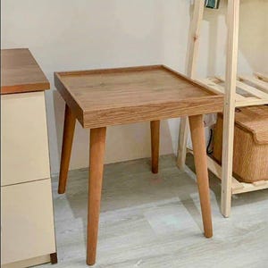 May include: A small, square wooden side table with a tray-like top and four tapered legs. The table is made of light brown wood and has a simple, mid-century modern design. The table is approximately 20 inches tall.