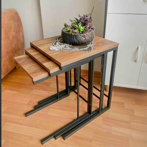 May include: Set of three nesting tables with a modern design. The tables have a dark metal frame and a wooden top. The top table has a decorative pot with plants. The tables are designed to slide under each other for space-saving.