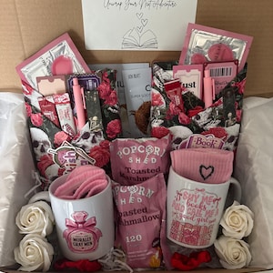 May include: A gift box containing two white ceramic mugs with pink graphics, two bags of toasted marshmallow popcorn, pink socks, skincare packets, and two wrapped items featuring a skull and rose pattern. A card reads Blind Date With A Book Box.