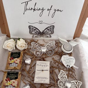 May include: A gift box containing a selection of items. Includes a card with "Thinking of you", silver butterflies, white roses, tea bags, heart-shaped sweets, a bracelet, and a candle holder.