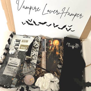 May include: A gift hamper labelled "Vampire Lover Hamper" containing themed items. Includes a face mask, socks with a bat design, a bookmark, a scrunchie, and other accessories. The box is decorated with bat and star motifs.