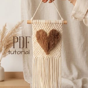 May include: Cream-colored macrame wall hanging with a fluffy brown heart design. The wall decor features a wooden dowel at the top and long fringe at the bottom. The text "PDF tutorial" is visible on the left side.