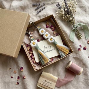 Handcrafted daisy macrame keychain gift for bridesmaid in a rustic box with proposal card, styled with dried petals, lavender and natural craft elements.