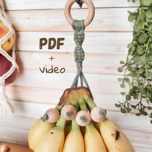 Könnte beinhalten: Ein handgefertigter Makramee-Bananenhalter mit Holzring und dekorativen Holzperlen. Der Halter hängt und zeigt reife Bananen. Der Text "PDF + video" ist sichtbar. Der Hintergrund zeigt eine Holzwand und Grün.