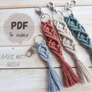 May include: Four macrame keychains in shades of blue, coral, cream, and teal. Each keychain features a woven design with a tassel and a silver-colored clasp and ring. The image also includes text that reads "PDF & video + BASIC KNOT GUIDE."