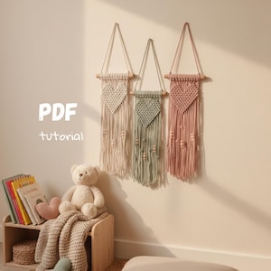 Könnte beinhalten: Drei Makramee-Wandbehänge in Creme, Salbeigrün und Altrosa, jeweils mit einem Herzdesign und Holzperlen. Die Aufhänger sind an Holzstäben befestigt. Der Text "PDF tutorial" ist an der Wand zu sehen.