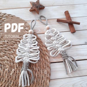 Könnte beinhalten: Zwei Makramee-Weihnachtsbaum-Ornamente in Grau- und Weißtönen, mit einer Holzperle und einer Schlaufe zum Aufhängen. Die Bäume werden auf einer gewebten Matte und einer Holzoberfläche präsentiert. Der Text "PDF TUTORIAL" ist sichtbar.