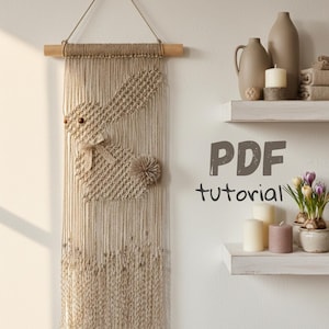 May include: A beige macrame wall hanging with a bunny design. The bunny has a textured body, a pom-pom tail, and a bow. The wall hanging is suspended from a wooden dowel. The text "PDF tutorial" is visible.