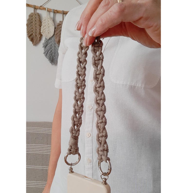 Peut inclure: Une sangle de t&eacute;l&eacute;phone en macram&eacute; beige avec des anneaux en m&eacute;tal argent&eacute;. La sangle est faite de cordon tiss&eacute; et nou&eacute;. L'&eacute;tui du t&eacute;l&eacute;phone est de couleur beige clair. Des plumes d&eacute;coratives sont suspendues en arri&egrave;re-plan.