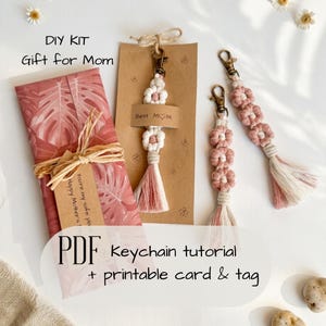 May include: A DIY kit with a macrame keychain tutorial. The kit includes a wrapped gift with a pink leaf pattern, a keychain with a white and pink flower design, and a tag that says "Best MOM". The text "PDF Keychain tutorial + printable card & tag" is also visible.