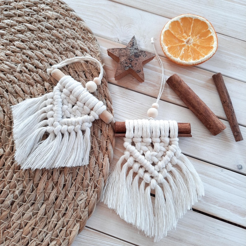 May include: Two macrame wall hangings with fringe, wooden beads, and a wooden dowel. A wooden star, dried orange slice, and cinnamon sticks are also present. The macrame is off-white, and the other items are brown.