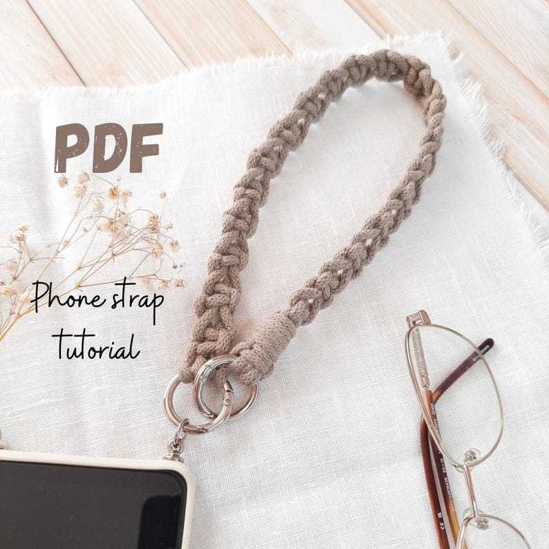 Peut inclure: Une lani&egrave;re de t&eacute;l&eacute;phone en macram&eacute; beige avec un fermoir argent&eacute; est attach&eacute;e &agrave; une coque de t&eacute;l&eacute;phone. La lani&egrave;re est boucl&eacute;e et porte le texte "PDF Phone strap Tutorial". La lani&egrave;re est pos&eacute;e sur une surface blanche.