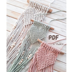 May include: Macrame wall hangings in cream, sage green, and pink, each with a heart design. The hangings are attached to wooden dowels and feature intricate knotting patterns. Some have wooden bead accents. The image includes the text "PDF + Video".