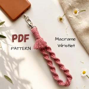 Puede incluir: Una pulsera de macramé rosa con un cierre plateado, una borla y un diseño en bucle. El texto "PDF PATTERN" es visible. Las margaritas y un teléfono están en el fondo.