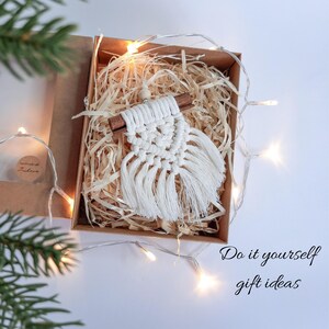 May include: A handmade macrame wall hanging in a gift box filled with straw. The white macrame is attached to a wooden dowel. The box is brown and the text "Do it yourself gift ideas" is written in cursive.