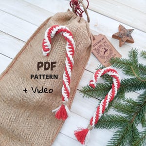 Puede incluir: Dos adornos de bastón de caramelo rojo y blanco con una bolsa de arpillera. La bolsa tiene el texto "PDF PATTERN + Video". Una estrella de madera y una etiqueta que dice "DELIVERED BY REINDEER MAIL 25TH DECEMBER" también están en la imagen.