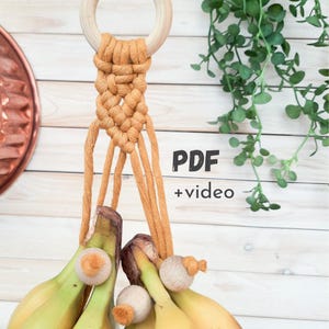 May include: A handmade macrame plant hanger in a warm tan color, featuring a wooden ring and a woven design. The hanger is holding a bunch of bananas. Text on the image reads "PDF + video".
