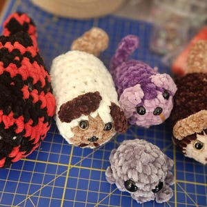 May include: A collection of handmade crochet animal toys in various colors. The toys include a red and black creature, a white dog-like figure with brown spots, a purple cat, a brown dog-like figure, and a gray creature. Each has black button eyes.