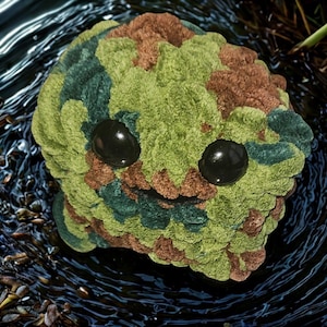 May include: A plush, round creature with a camouflage pattern in shades of green, brown, and blue. It has two large, black, glossy eyes and a stitched smile. The creature is set against a dark, watery background.