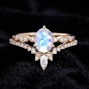 May include: A gold ring set featuring a central oval moonstone with a blue iridescent glow, flanked by small round diamonds on the band. A matching chevron-shaped band sits below, adorned with small round diamonds and a single marquise diamond.