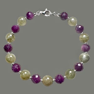 May include: A beaded bracelet featuring alternating faceted, round beads. The bracelet has a silver clasp and is composed of purple and olive-green gemstones. The beads are strung together with a silver chain.