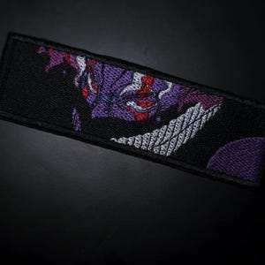 Peut inclure: Écusson rectangulaire noir brodé avec un motif violet, blanc et rouge. Le motif représente un visage ou un masque stylisé. L'écusson est probablement destiné aux vêtements ou aux accessoires.