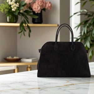 May include: A black suede handbag with two rounded handles. The bag has a structured shape with a top zipper closure and a small pocket on the front. The bag is on a white marble surface.