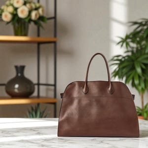 May include: A brown leather handbag with two top handles. The bag has a rounded top and a rectangular base. The bag is on a marble surface. A vase of white roses and a green plant are in the background.