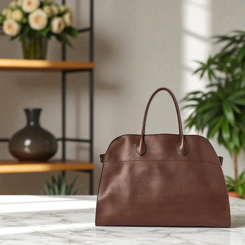 May include: A brown leather handbag with two top handles. The bag has a rounded top and a rectangular base. The bag is on a marble surface. A vase of white roses and a green plant are in the background.
