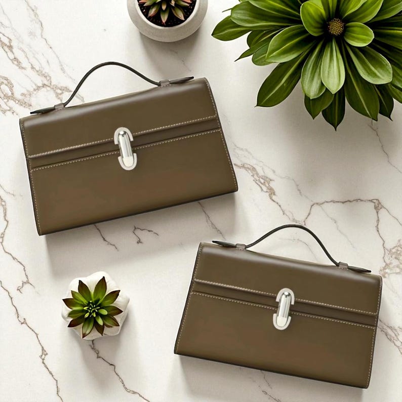 Op de afbeelding: Twee olijfgroene handtassen met zilveren sluitingen en zwarte handvatten worden getoond op een marmeren oppervlak. De tassen hebben een rechthoekige vorm met een gestikt ontwerp. Decoratieve planten in kleine potten zijn rond de tassen geplaatst.