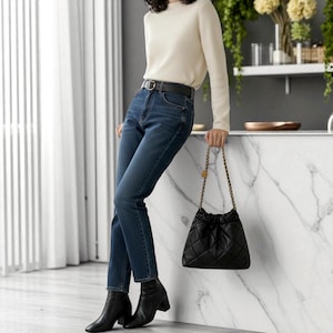 Puede incluir: Una mujer con un su&eacute;ter crema de manga larga, vaqueros azul oscuro y botines negros. Sostiene un bolso negro acolchado con cord&oacute;n y correa de cadena dorada. Un cintur&oacute;n negro con hebilla dorada en la cintura.