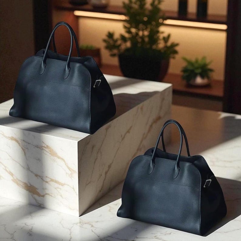May include: Two navy blue leather handbags with top handles and silver-tone hardware. The bags are displayed on a marble-effect surface, with a plant in the background. The bags have a structured shape and a minimalist design.