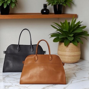 May include: Two leather handbags. One is black, the other is tan. Both have a rounded shape, top handles, and a horizontal seam across the middle. The bags are on a marble surface, with plants in the background.