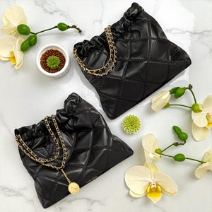 Puede incluir: Dos bolsos negros acolchados con cord&oacute;n y correas de cadena doradas. Las bolsas tienen un patr&oacute;n de diamantes y se exhiben sobre una superficie de m&aacute;rmol, junto con orqu&iacute;deas blancas y una peque&ntilde;a planta suculenta.