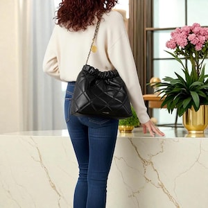 Puede incluir: Bolso negro de cuero acolchado con cord&oacute;n y correa de cadena dorada. El bolso tiene un patr&oacute;n de diamantes y una parte superior fruncida. El bolso se lleva al hombro. El fondo incluye un su&eacute;ter crema, vaqueros azules y una encimera de m&aacute;rmol.