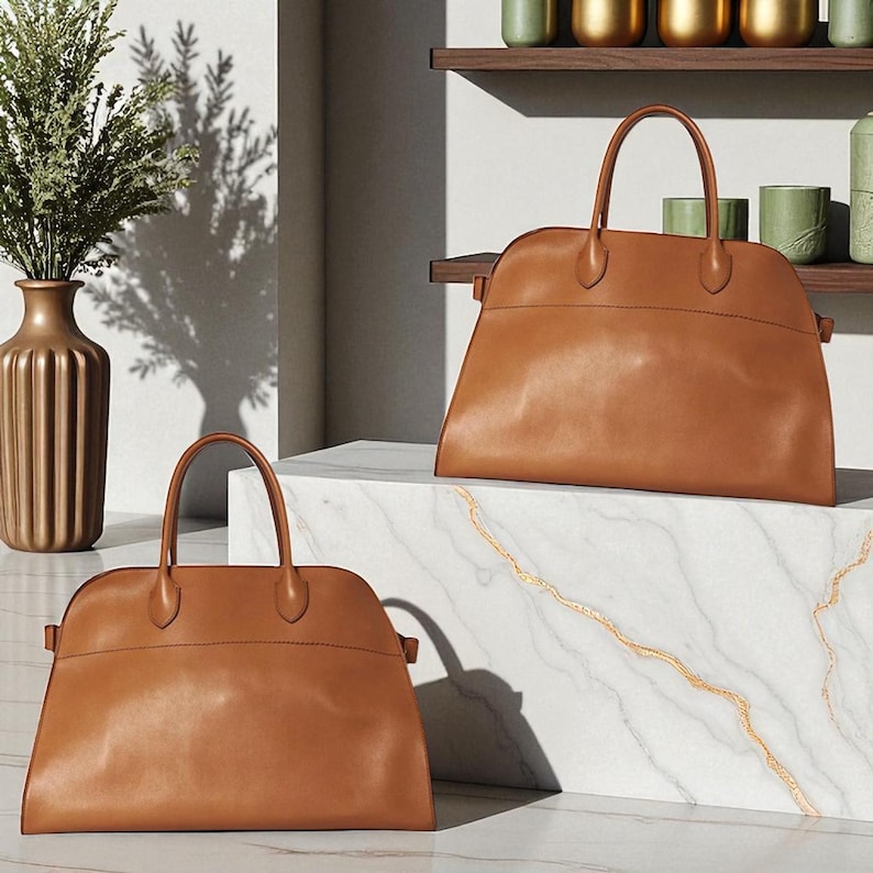 May include: Two tan leather handbags with top handles. The bags have a structured shape and a smooth finish. One bag is on a white marble surface, and the other is on a similar surface. A gold vase and greenery are in the background.