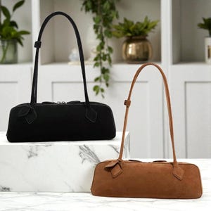 Può includere: Due borse a tracolla in camoscio, una nera e una marrone. Entrambe hanno cinghie lunghe e sottili e una forma rettangolare con un piccolo dettaglio decorativo sul davanti. Le borse sono esposte su una superficie di marmo bianco.