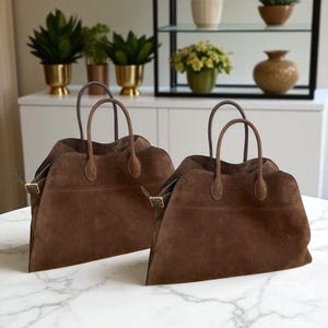 May include: Two brown suede tote bags with top handles and gold-coloured buckles. The bags are rectangular with a trapezoidal shape and are displayed on a white marble surface. The bags have a classic, minimalist design.